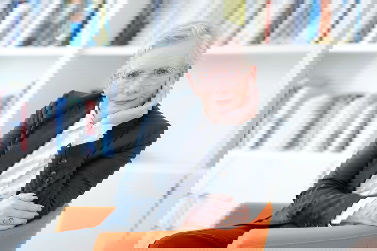 Eine Frau mit sehr hellen Haaren und Brille und dunklem Jackett sitzt in einem orangenen Sessel vor Bücherregalen.