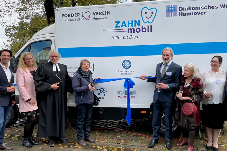 Vor einem weiß-blauen Kastenwagen, auf dem "Zahnmobil" steht, stehen mehrere lachende Menschen. Zwei halten eine blaue Schleife.