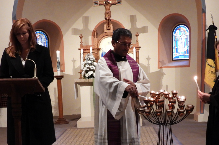 Eine Person steht an einem Pult in einer Kirche, daneben zündet ein Mann Kerzen an, daneben steht eine Frau und zündet ebenfalls Kerzen an.