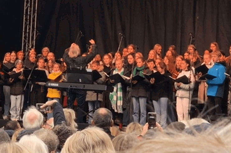 Aus einer Menschenmenge heraus sieht man eine Bühne, auf der Menschen mit Gesangmappen stehen.