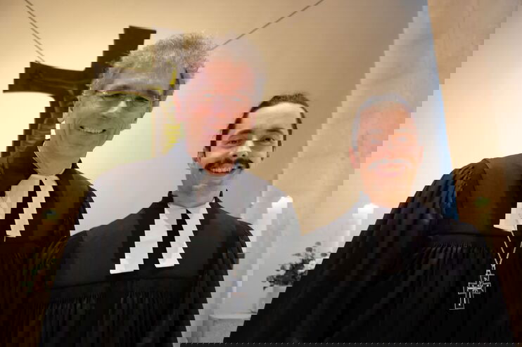 Zwei Pastoren in schwarzen Talaren stehen vor einem Kreuz. Links und rechts sind weiße Altarkerzen zu sehen