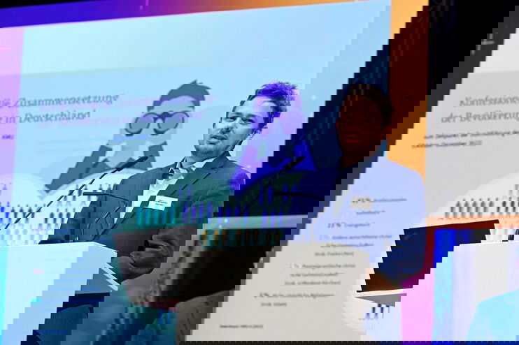 Ein junger Mann spricht an einem Redepult, hinter ihm ist auf einer Leinwand eine Präsentation zu sehen.