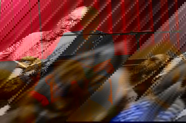 Ein Mann im grauen Sakko sitzt hinter einem Mikrofon und liest Kindern etwas vor.