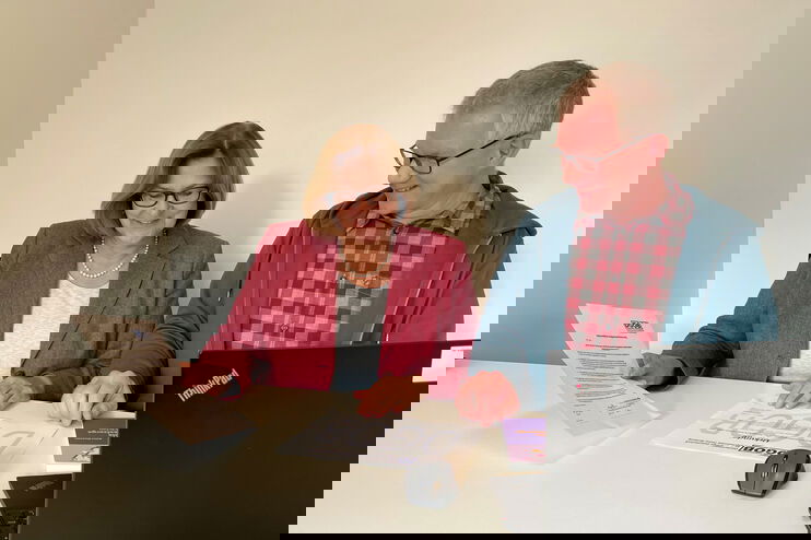 Ein Mann und eine Frau sitzen an einem Tisch, darauf steht ein Laptop. Die Frau hält ein Blatt in der Hamd.