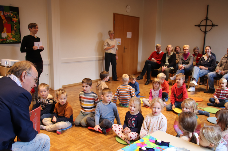 Links sitzt ein Mann mit Jeans und blauem Jackett und Halbglatze. Vor ihm sitzen Kinder, teils auf Kissen. Rechts an der Wand hängt ein großes Kreuz.