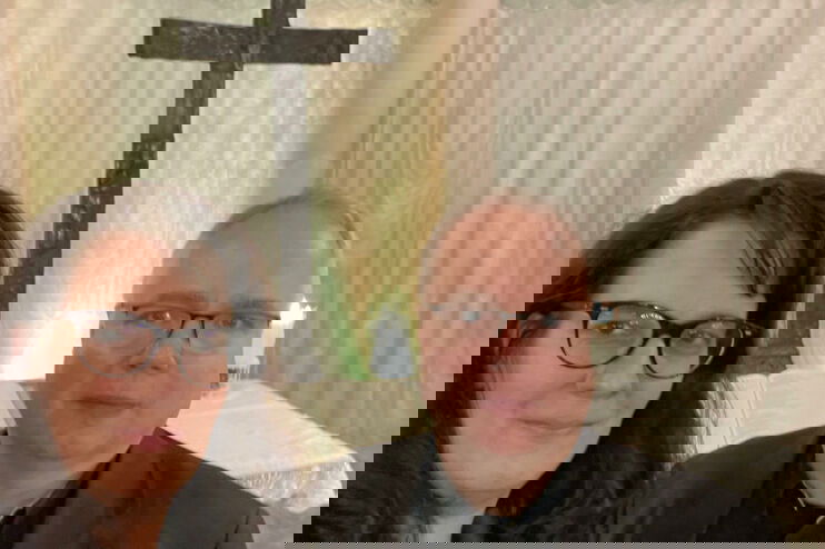 eine weiblich und männlich gelesene Person stehen vor einem Altar und schauen in die Kamera.