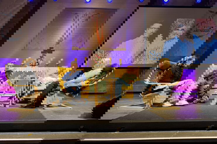 Vier Personen sitzen in Stühlen auf einer kleinen Bühne vor einem Altar. Rechts im Hintergrund ist eine Leinwand aufgebaut, auf der zwei Personen zu sehen sind: Steffen Hallaschka und Olivia Jones.