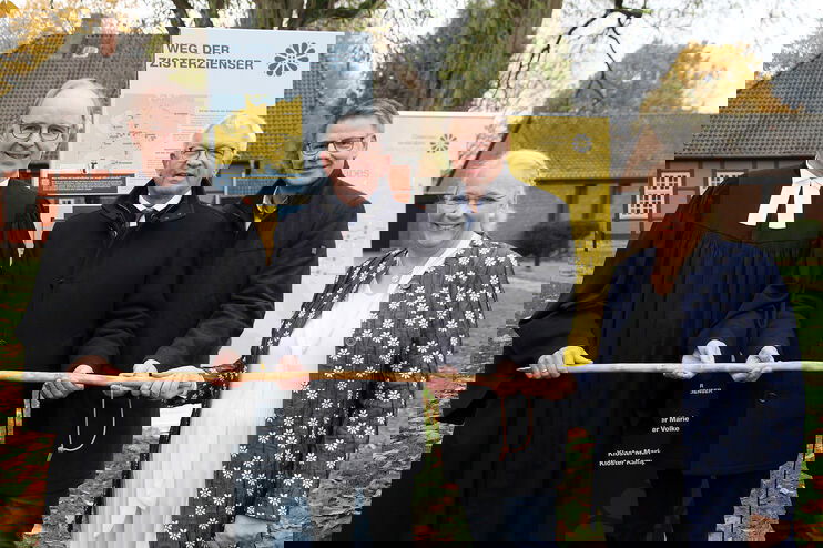 Drei Männer und eine Frau halten einen Wanderstab vor einer Infosäule.