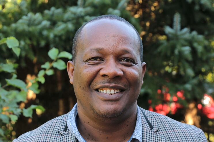 Ein schwarzer Mann steht lächelnd im Grünen. Er trägt ein graues Sakko und ein hellblaues Hemd.