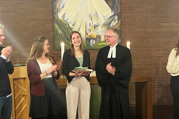 Mehrere Personen stehen nebeneinander, alle bis auf eine klatschen. Diese hält eine Mappe in Händen. Eine Person trägt einen Talar.