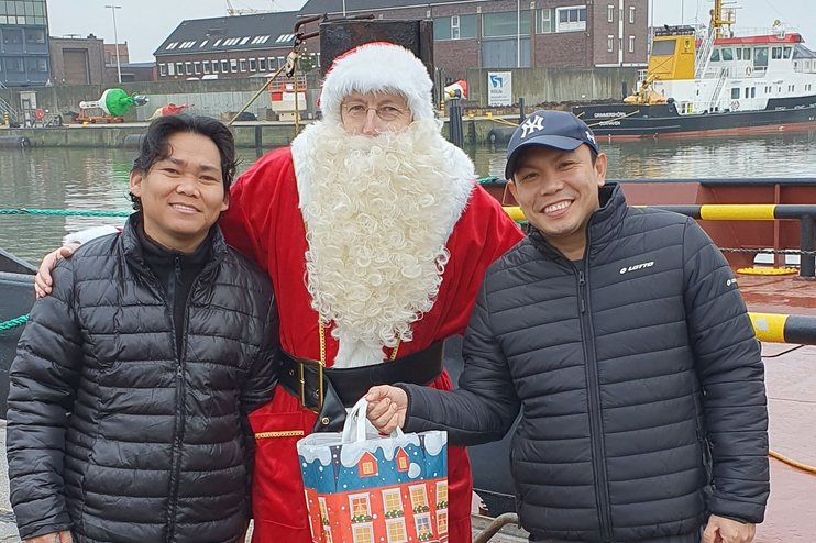 Zwei Männer mit Winterjacken halten Geschenktüten und stehen links und recht von einem Weihnachtsmann.