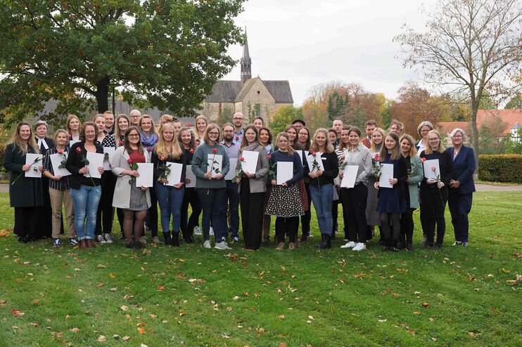 Viele eher jüngere Menschen stehen zusammen auf einem Rasen und halten Urkunden in Händen. Im Hintergrund ragt spitz eine Kirche auf.