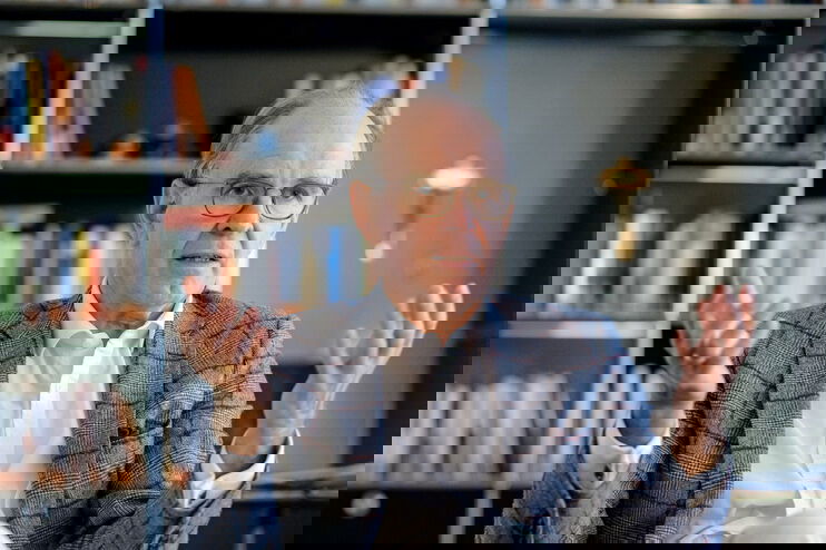 Ein älterer Mann mit weißem Hemd und grauem Sakko sitzt vor einer Bücherwand und spricht gestenreich.