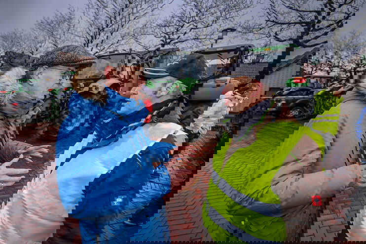 Zwei Männer, einer mit blauer, einer mit gelber Warnweste über einer braunen Jacke, beide mit Mützen, stehen sich gegenüber, der links gestikuliert. Im Hintergrund stehen Traktoren.