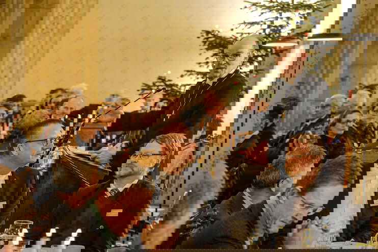 Viele Menschen sitzen an mehreren Tischen zusammen, einer steht zwischen ihnen, mit schwarzer Robe und Kreuz um den Hals. Im Hintergrund ein Weihnachtsbaum.