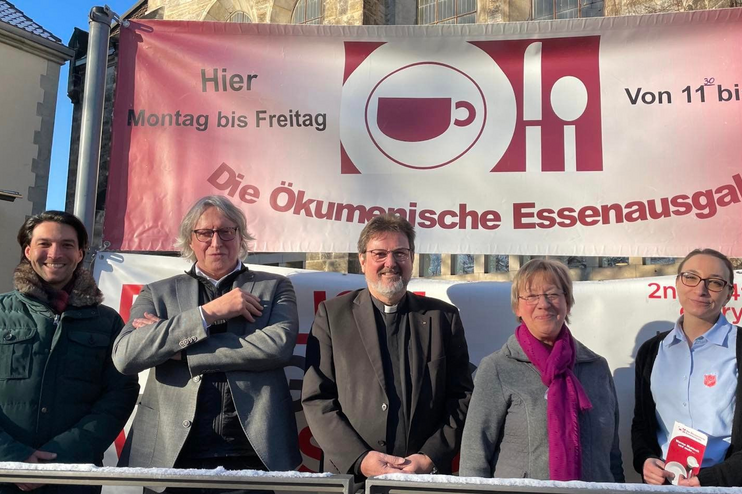 Fünf Menschen stehen in Reihe vor einem roten Plakat, auf dem steht: Ökumenische Essensausgabe, Montag bis Freitag, 11 bis 13 Uhr. 