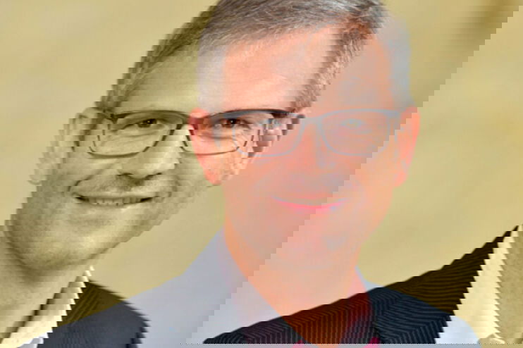 Ein Mann mit kurzen grauen Haaren und Brille, rosa Hemd und dunklem Jackett darüber, lächelt.