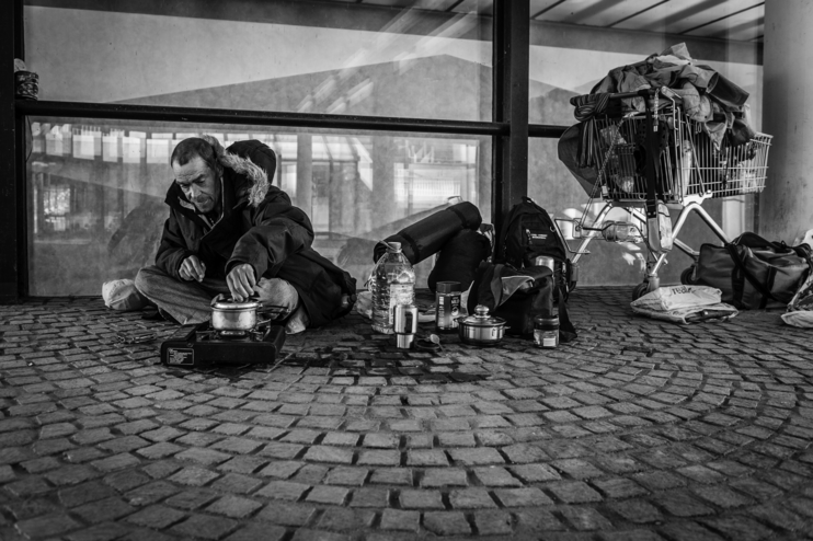 Ein schwarz-weiß-Bild zeigt einen wohl obdachlosen Mann, der mit einige Habseligkeiten auf der Straße vor einem Laden sitzt.