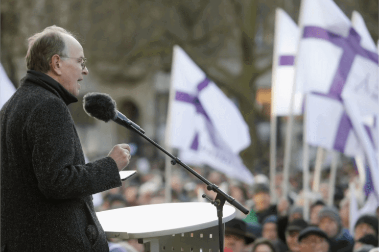 Eine männlich zu lesende Person spricht in ein Mikrofon. Im Hintergrund sind Menschen zu erkennen, die Fahnen mit violetten Kreuzen schwenken.