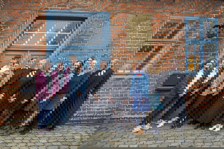 Gruppe von Menschen vor einem Backsteingebäude mit grüner Türe. 12 Personen, zwei davon tragen einen Talar.