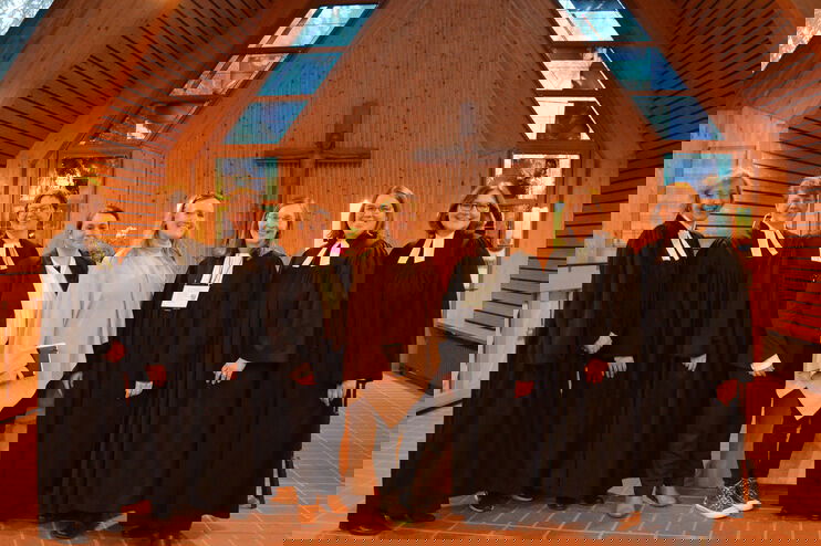 In einer mit Holz ausgekleideten Kappe stehen acht Frauen in einer Reihe. Jeweils die ersten drei von links und die ersten drei von rechts tragen Talar. In der Mitte stehen zwei Frauen, die eine trägt einen schwarzen Hosenanzug, die andere einen hellen Pulli und eine schwarze Hose. Alle lächeln in die Kamera. 