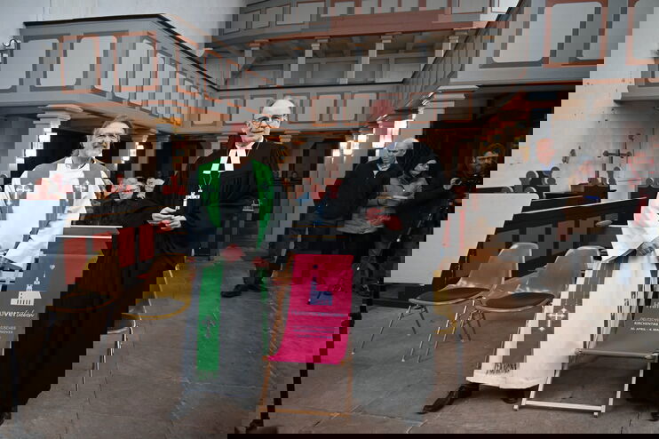 Zwei Männer,, einer im weißen, einer im schwarzen Talar, stehen links und recht eines rot-rosa Liegestuhls. Im Hintergrund eine Kirche, in deren Bänke Menschen sitzen.