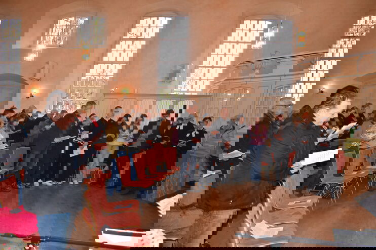 Mehrere Menschen stehen in einem Halbkreis vor Stühlen in einer Kirche mit hohen Fenstern. Manche tragen Talar.