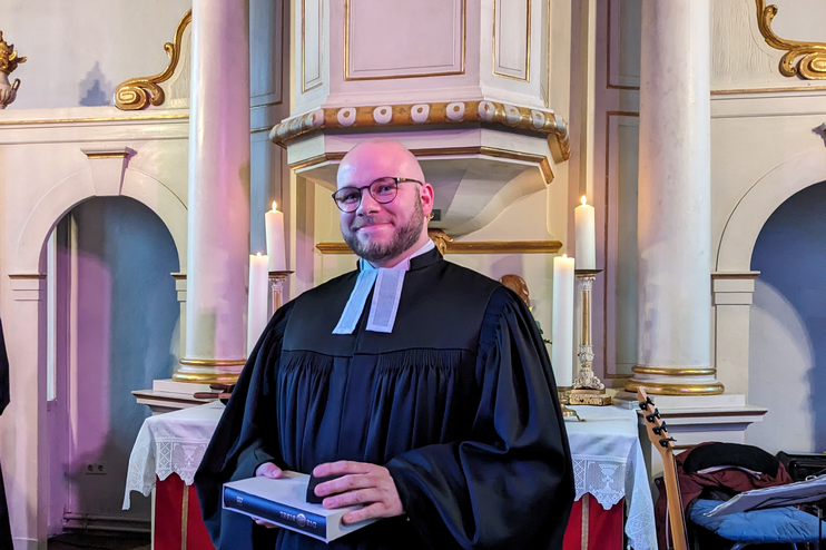 Ein Mann in Talar steht in einer hellen Kirche vor dem Altar mit rotem Parament. Er hält ein Buch in den Händen.
