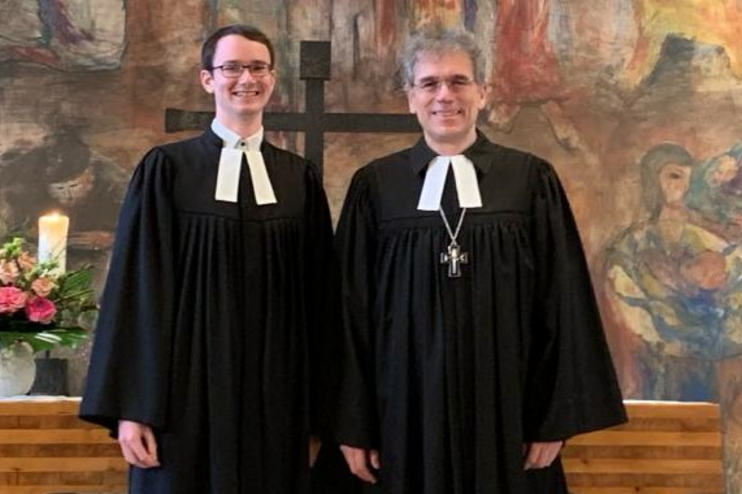 Zwei Männer in Talar stehen vor einem hölzernen Altar und lächeln in die Kamera. Im Hintergrund ist ein buntes Wandgemälde zu sehen.