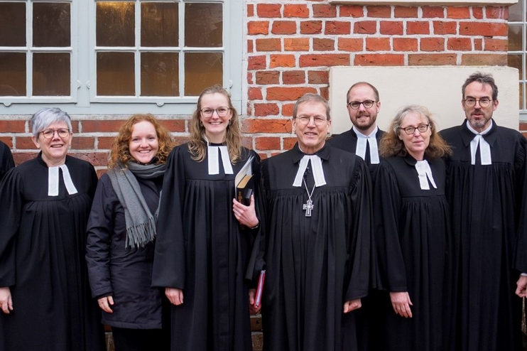 Eine Gruppe von sieben Menschen in Talar steht vor einer Backsteinmauer. Alle lächeln in die Kamera.