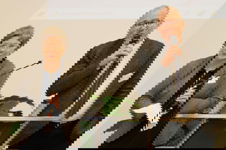 Eine ältere Frau und ein älterer Mann stehen an einem Pult und sprechen in ein Mikrofon.