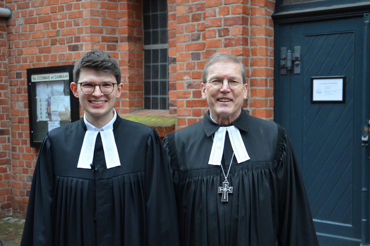 Zwei Männer tragen Talar, lächeln in die Kamera. Sie stehen vor einem Backsteingebäude mit dunkler Holztüre.