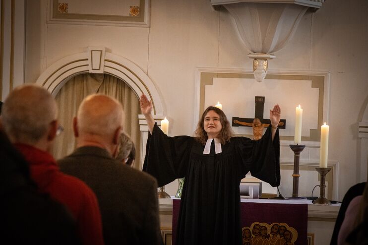 Eine Frau im Talar spendet einer Gemeinde den Segen. Im Vordergrund sind die Rücken von zwei Menschen zu sehen. Hinter der Frau, die den Segen spendet, ist ein Altar mit rotem Parament zu sehen.