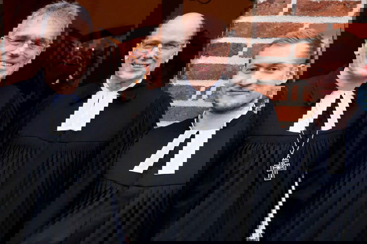 Drei Männer in Talar stehen vor einer Kirche und lächeln in die Kamera.