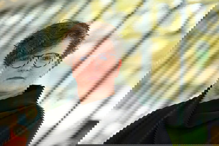 Eine als Frau lesbare Person mit kurzen Haaren vor einer Glaswand.