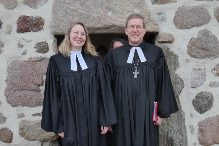 Vor einer gemauerten Fassade treten aus einer Türe eine junge frau mit blonden Haaren und Brille und neben ihr ein Mann mit kurzen braunen Haaren und Brille. Beide tragen Talar.
