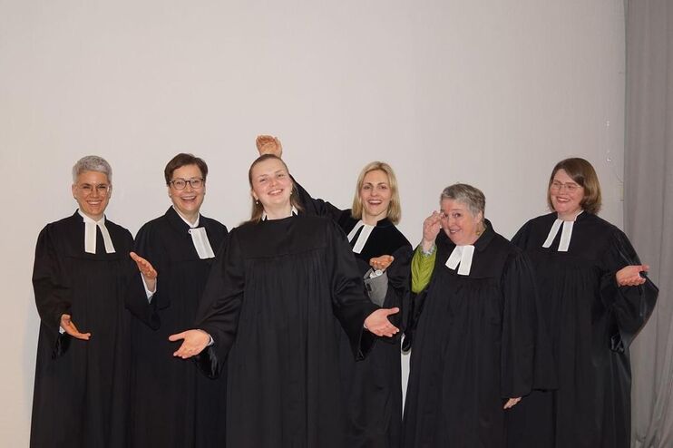 Sechs Personen in Talaren stehen zusammen und machen Gesten mit den Handflächen nach oben, nach dem Motto: "Hier sind wir"