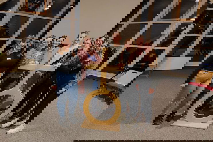 Sechs weiblich gelesene Personen und eine männliche, alle jung, stehen um ein Holzkreuz auf einer Kugel.