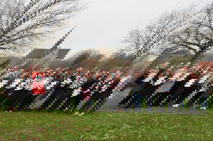 Eine Gruppe von Menschen hält Urkunden und rote Rosen in den Händen. Sie stehen auf einer Wiese vor einer Kirche und sind festlich gekleidet.