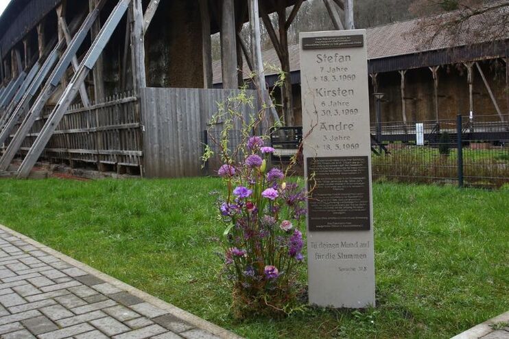 Eine steinerne Stele mit Namen drauf steht auf einer grünen Wiese. Im Hintergrund sind Gebäude aus Holz zu erkennen.