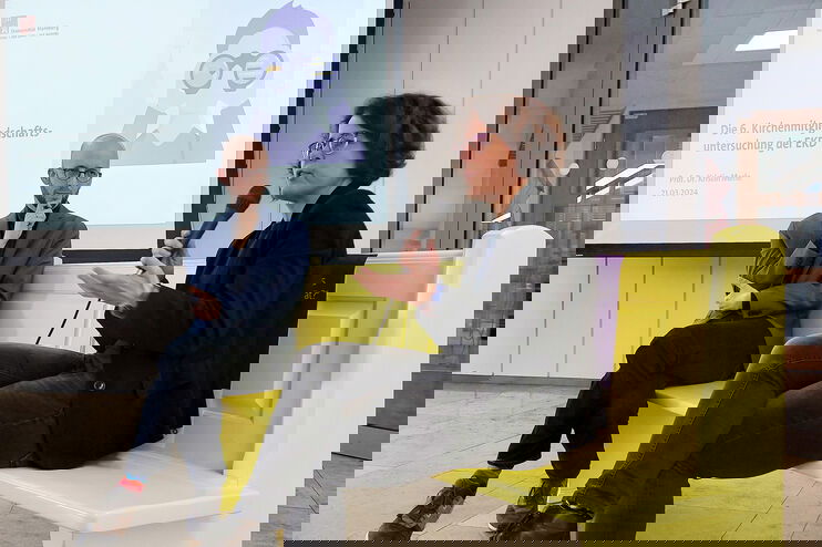 Eine als Frau lesbare Person sitzt auf einer gelben Bank und spricht in ein Mikrofon. Eine als Mann lesbare Person sitzt daneben und hört zu.