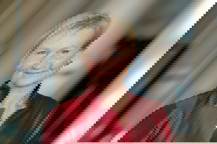 Eine weiblich gelesene Person mit kurzen, blonden Haaren und in satin-rotem Oberteil lächelt fröhlich. 