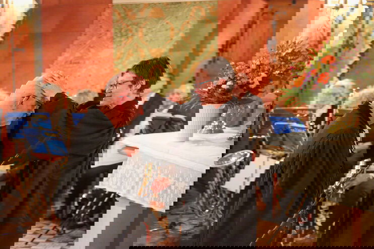 Eine weiblich gelesene Person legt einer männlich gelesenen Person die Hände auf die quasi-Glatze. Beide Tragen Talar. Im Hintergrund sitzen Menschen an Notenständern im Altarraum einer Kirche.