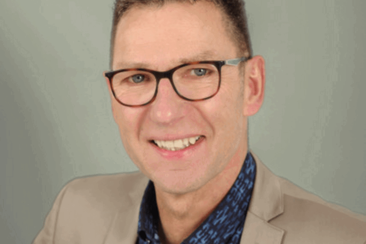 Eine als Mann lesbare Person mittleren Alters mit kurzen Haaren, Brille, dunkelblau gemustertem Hemd und hellbraunem Sakko.