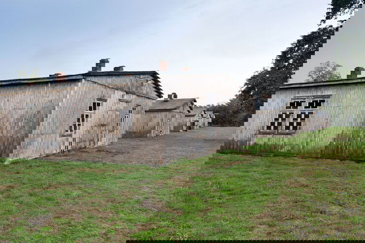Einfache Bretter bilden eine Holzhütte mit wenig Fenstern auf einem stoppeligen Rasen.