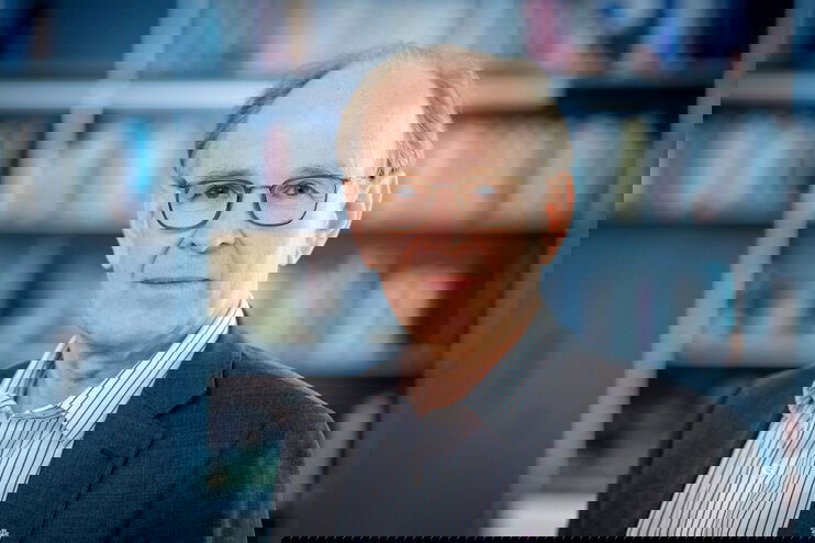 Eine als Mann lesbare Person mit Brille, Hemd und Sakko steht vor einer Bücherwand und schaut in die Kamera.