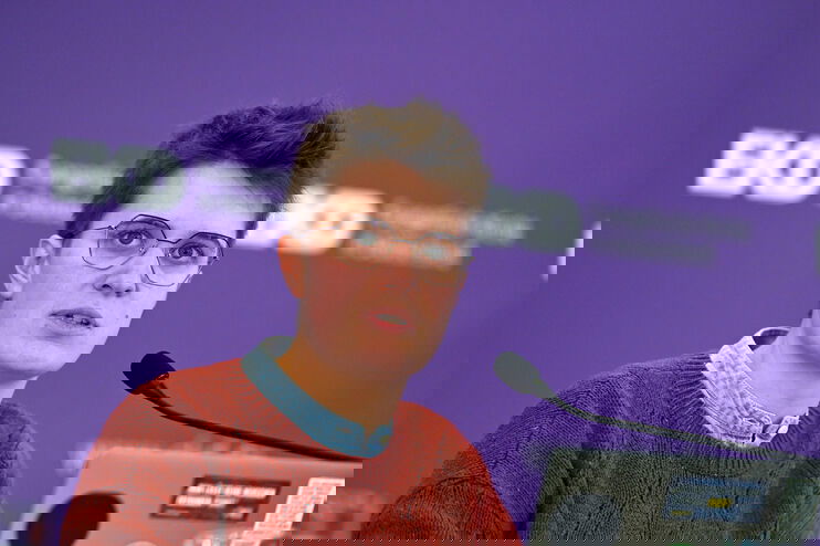 Eine junge als Frau lesbare Person mit Kurzhaarschnitt, Brille und rotem Strickpullover spricht in ein Mikrophon.