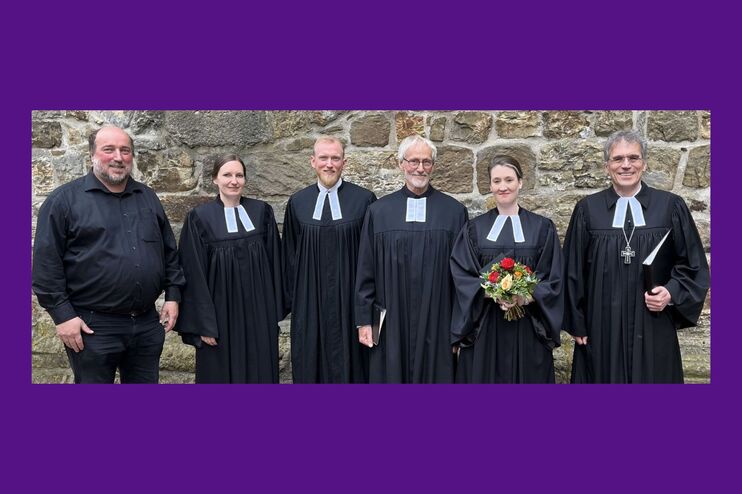 Sechs Personen stehen nebeneinander vor einer steinernen Wand. Fünf tragen Talar, einer ein dunkles Hemd. Eine weiblich gelesene Person hält einen Blumenstrauß in Händen.