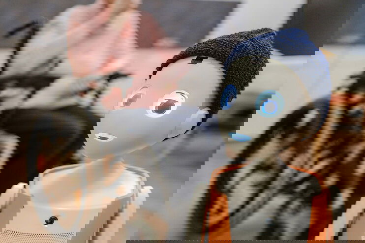 Eine weiblich gelesene Person sitzt im Rollstuhl. Vor ihr steht ein Roboter, der einem Jungen nachempfunden ist, Mund und Augen leuchten blau, mit orangener Latzhose und blauer Mütze.