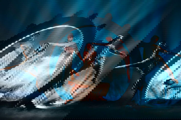 Eine als Frau lesbare Person singt in einem beleuchteten Kreis, umgeben von Tänzern.